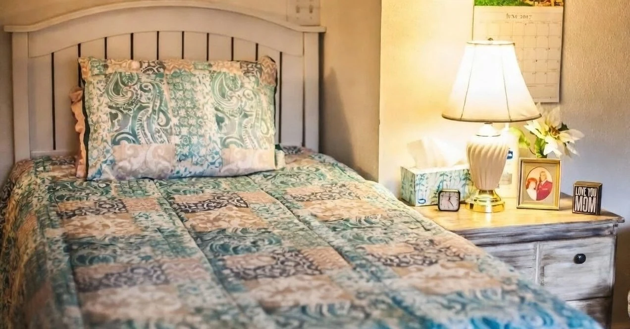 Furnished bedroom with a patterned quilt, pale headboard and a lamp on the bedside cabinet.